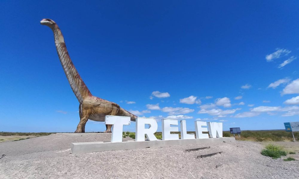 Cartel de Trelew con el monumento al dinosaurio más grande del mundo detrás. 